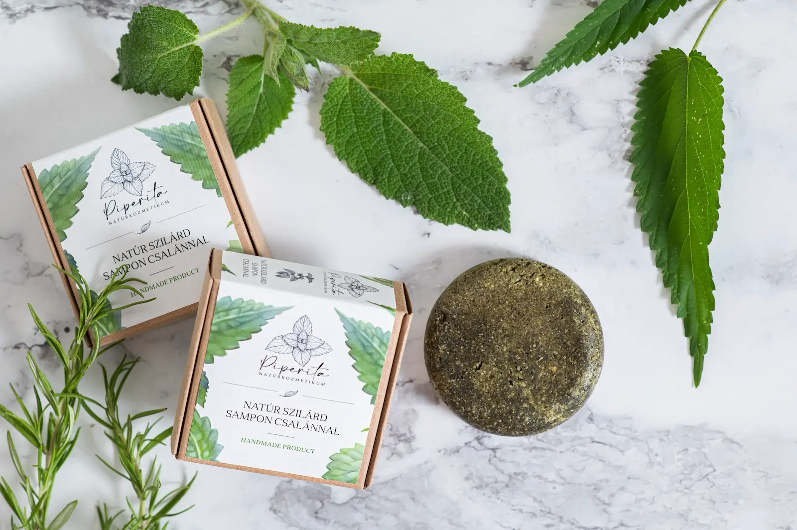 Piperita natúr szilárd sampon csalánnal, és két újrahasznosítható papírdobozos csomagolásban, friss csalánlevelekkel és rozmaringággal, márványfelületen elrendezve.