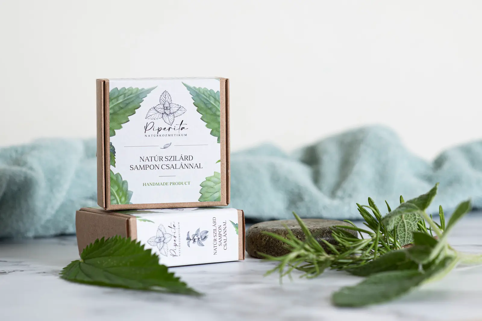 Piperita natúr szilárd sampon csalánnal, két újrahasznosítható papírdobozban, rozmaring- és csalánlevelekkel, világos háttérrel, természetes környezetben.