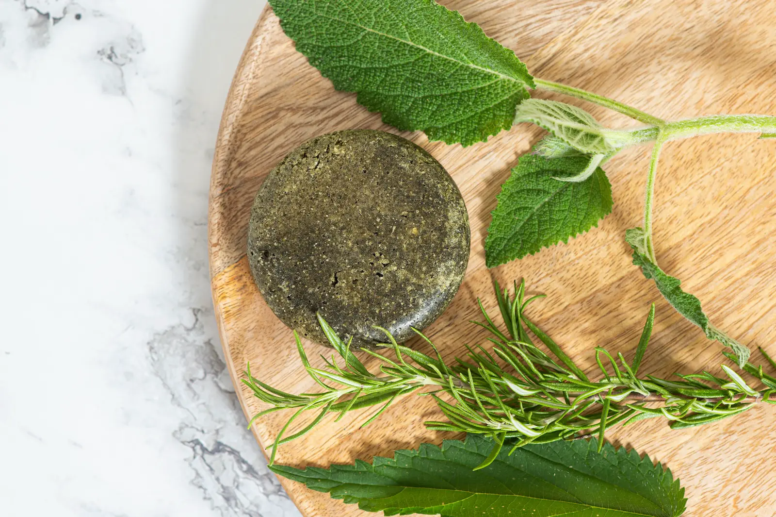 Zöldes-barna színű szilárd sampon korong csalán- és rozmaringlevelekkel egy világos fa tálcán, természetes fényben.