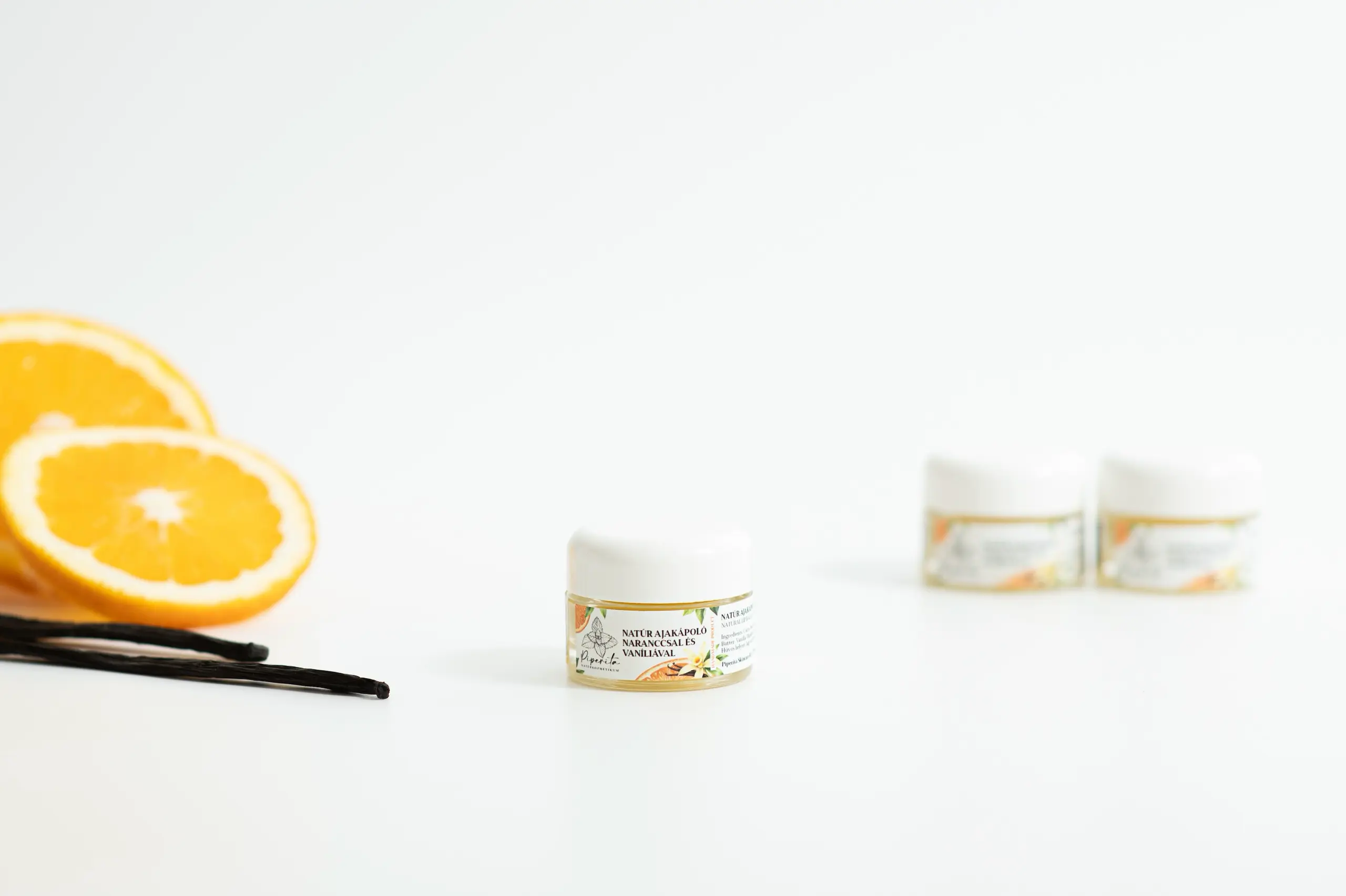 Natúr ajakápoló narancs és vanília összetevőkkel, friss narancsszeletek és vaníliarúd mellett világos háttéren