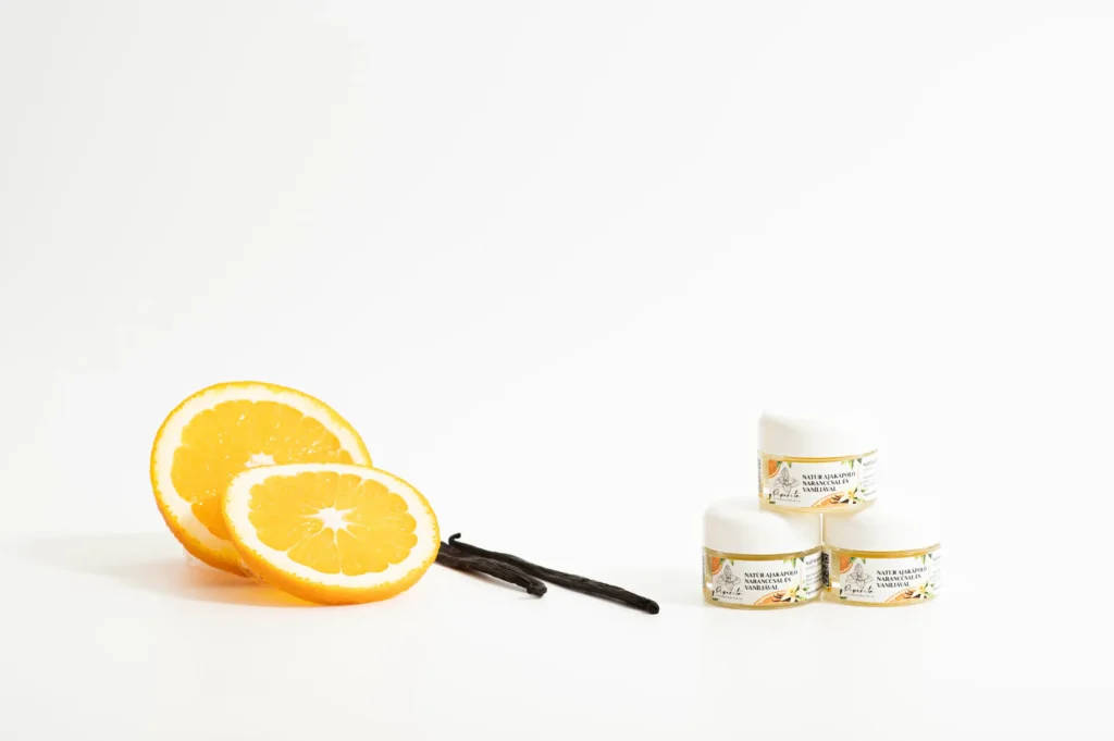Natúr ajakápolók narancs és vanília összetevőkkel, narancsszeletek és vaníliarúd mellett világos háttéren