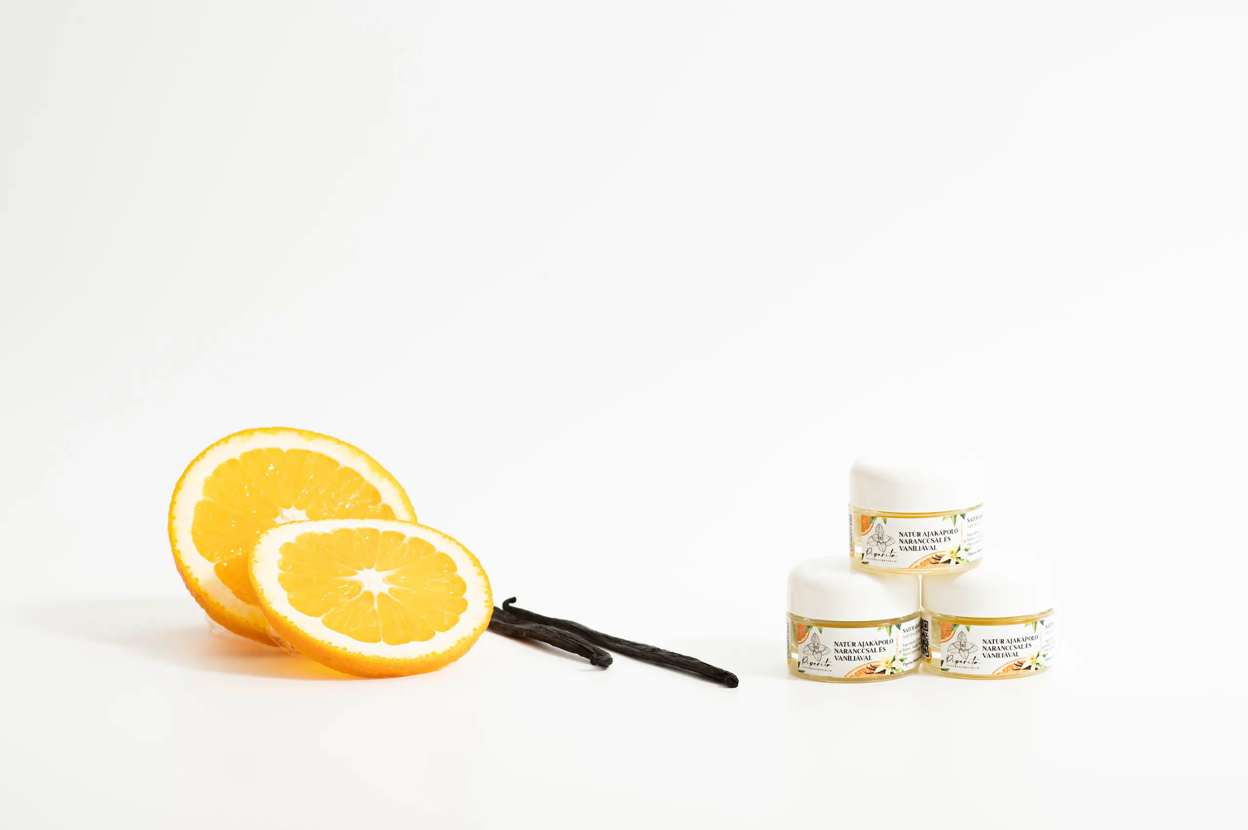 Natúr ajakápolók narancs és vanília összetevőkkel, narancsszeletek és vaníliarúd mellett világos háttéren